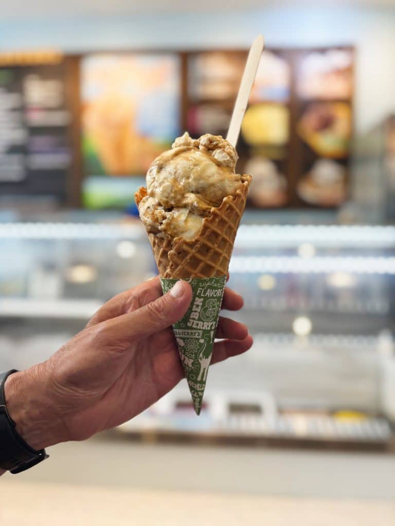 Eine Hand hält eine Waffeltüte mit zwei Kugeln Ben & Jerry's Eiscreme in einem Restaurant in Montreal, ergänzt durch einen kleinen Holzlöffel.