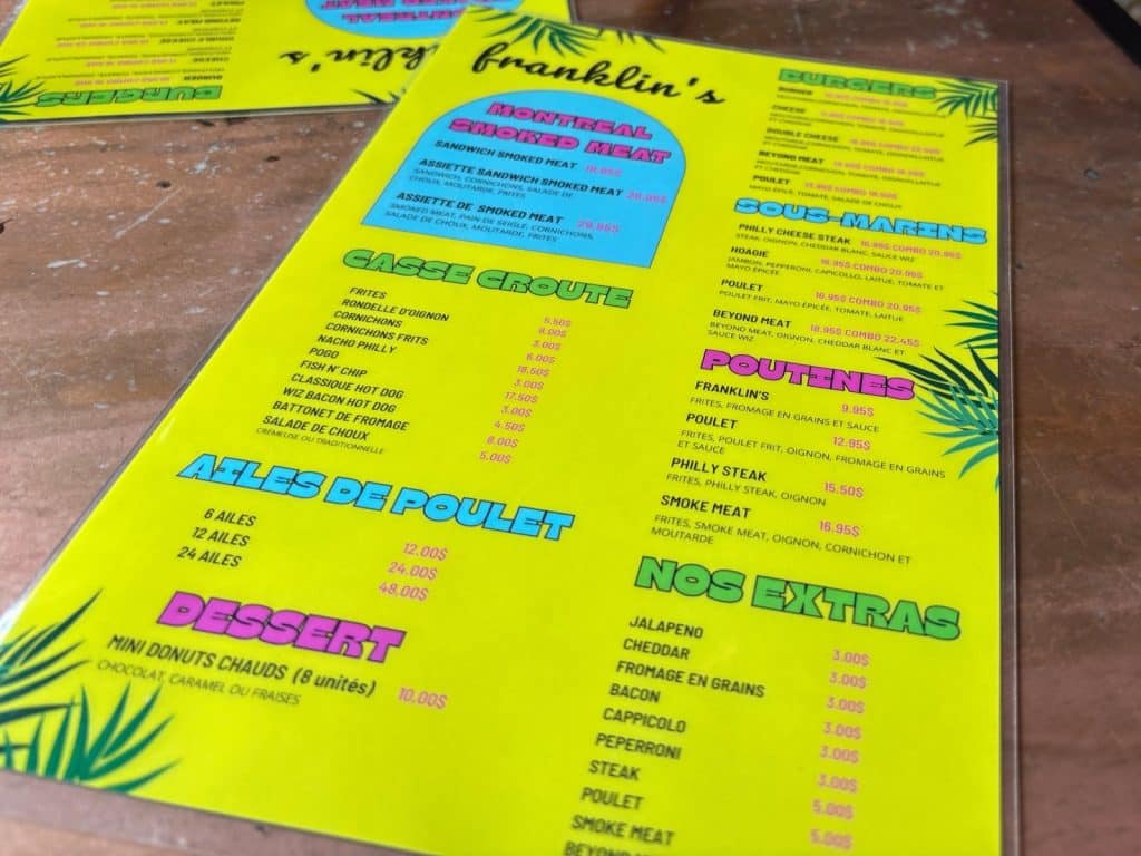 Restauranttipp Montreal: Eine neongelbe Speisekarte des Restaurants Franklin's in Montreal zeigt verschiedene Kategorien wie Montreal Smoked Meat, Casse-Croûte, Poutines und Burger mit den dazugehörigen Preisen in kanadischen Dollar. Die Karte ist mit blauen und pinken Akzenten sowie Palmenblatt-Grafiken gestaltet und liegt auf einem Holztisch.