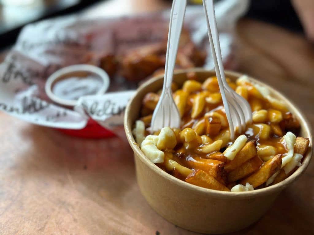 Eine Nahaufnahme einer Portion Poutine von Franklin's in Montreal, serviert in einer Pappschale mit Pommes frites, Käsequark, brauner Bratensauce und zwei weißen Plastikgabeln.