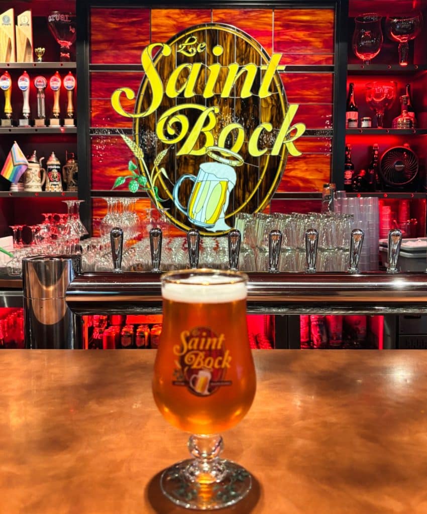 Ein Glas frisch gezapftes Bier steht auf dem Tresen der Brasserie Le Saint-Bock in Montreal, im Hintergrund ist das beleuchtete Logo der Bar sowie eine Reihe von Zapfhähnen zu sehen.