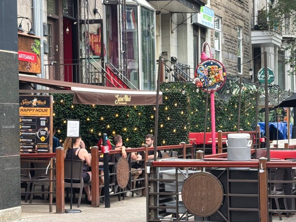 Der Außenbereich der Brasserie Le Saint-Bock in Montreal zeigt Gäste, die an Tischen auf einer von einer Hecke mit Lichterketten gesäumten Terrasse sitzen, während im Hintergrund die städtische Architektur und bunte Schilder zu sehen sind.