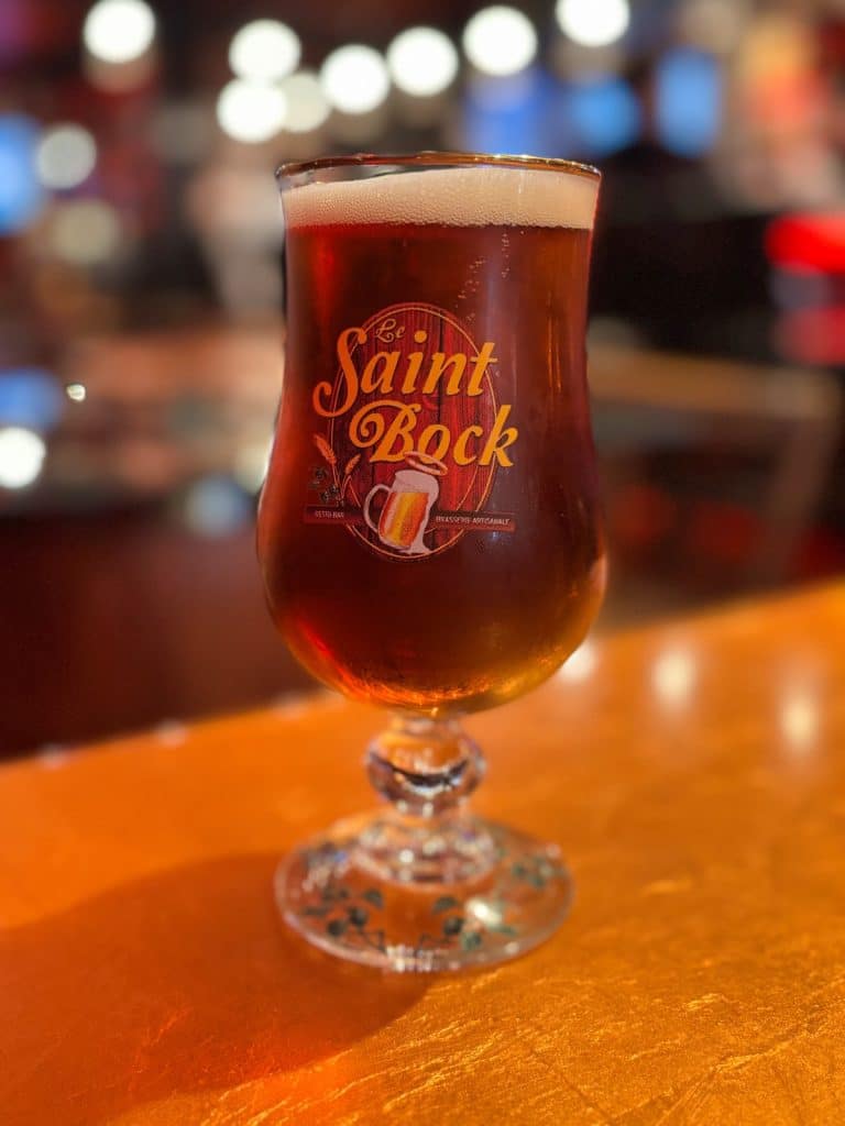 Ein Glas dunkles Bier der Brauerei Le Saint-Bock in Montreal steht auf einer beleuchteten Theke vor einem unscharfen Hintergrund mit Lichtern. Das geschwungene Glas zeigt das Logo der Brasserie Artisanale und ist mit einer feinen Schaumkrone gefüllt.