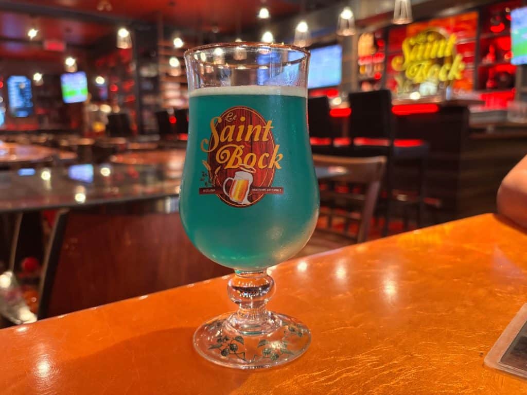 Ein Glas mit leuchtend blauem Bier steht auf einem hölzernen Tresen in der Brasserie Artisanale Le Saint-Bock in Montreal, während im Hintergrund die stimmungsvoll beleuchtete Bar unscharf zu sehen ist.