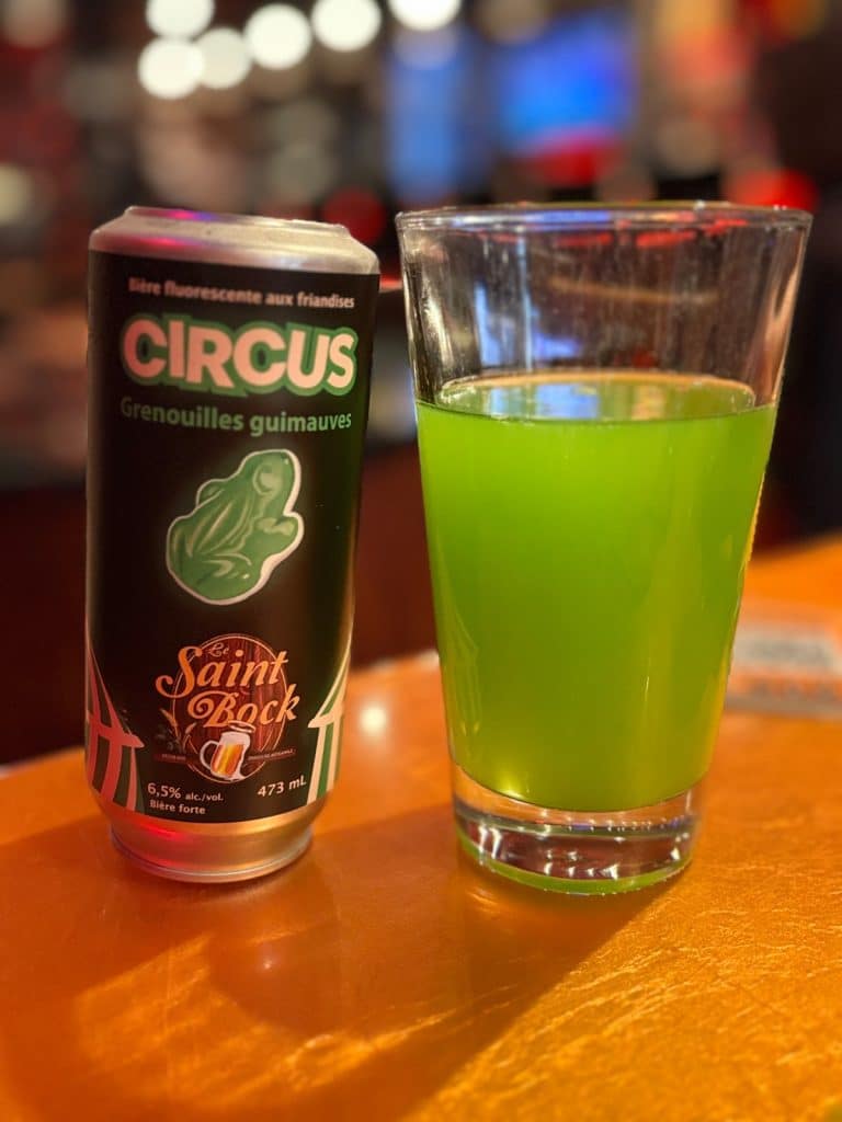 Eine schwarze Dose des Bieres „Circus Grenouilles guimauves“ von der Montrealer Brauerei Le Saint-Bock steht neben einem Glas, das mit einer leuchtend grünen, trüben Bier gefüllt ist.