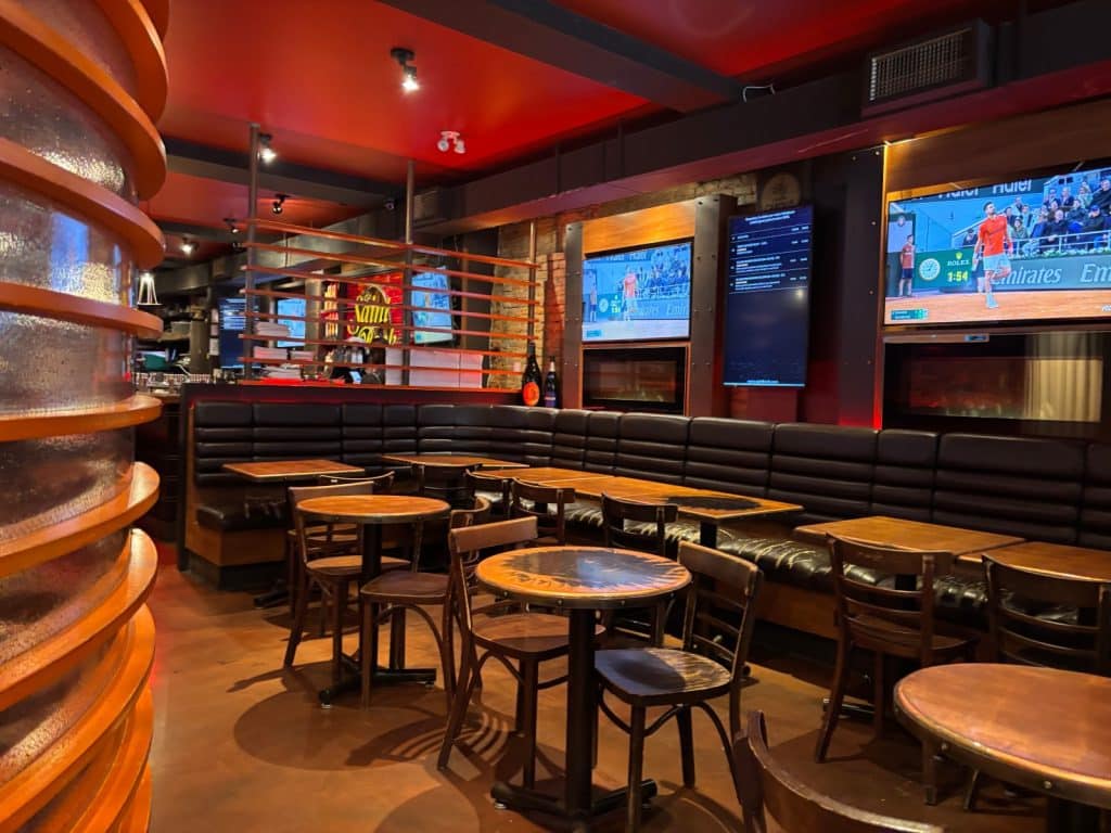 Innenansicht der Bar Le Saint-Bock in Montreal mit dunklen Lederbänken, Holztischen und mehreren Bildschirmen an den Wänden unter einer roten Decke.