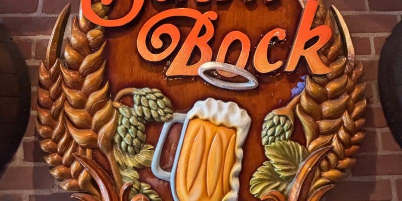 Ein kunstvoll geschnitztes Holzschild des Montrealer Pubs „Le Saint Bock“ zeigt den Namen in geschwungener oranger Schrift, umrahmt von Getreideähren, Hopfen und einem schäumenden Bierkrug mit Heiligenschein vor einer Backsteinwand.