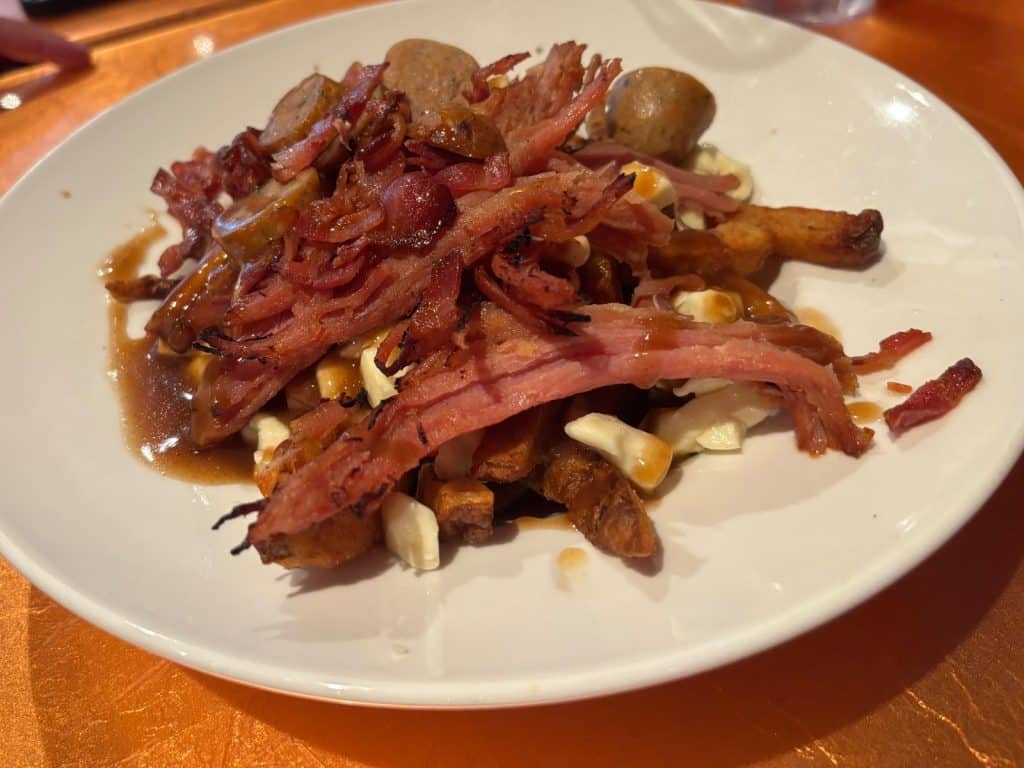Eine Portion Poutine auf einem weißen Teller, bestehend aus Pommes frites, Käsebruch und Bratensoße, die großzügig mit knusprigen Speckstreifen und Wurstscheiben belegt ist.