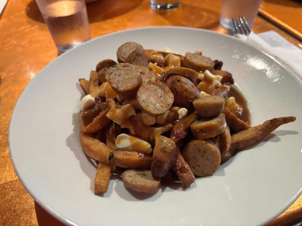 Eine Portion Poutine, bestehend aus Pommes frites, Käse-Curds und brauner Bratensauce, garniert mit zahlreichen Scheiben gebratener Wurst auf einem weißen Teller.