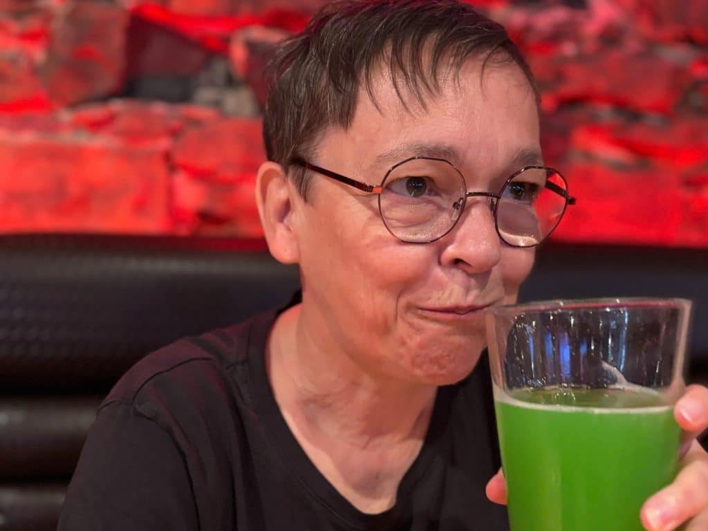 Eine Person mit Brille lächelt leicht, während sie in der Brasserie Le Saint-Bock in Montreal ein Glas mit leuchtend grünem Bier in der Hand hält.