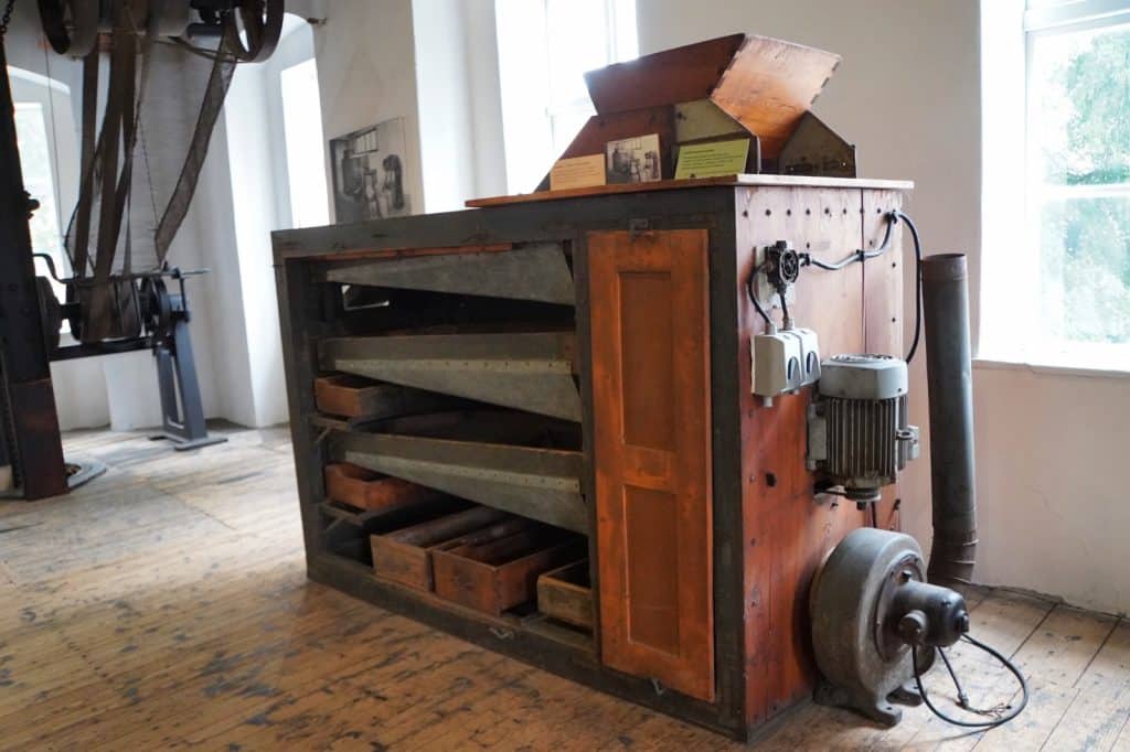 Eine alte, hölzerne Maschine im Hopfenmuseum in Saaz verfügt über mehrere schräge Metallebenen und Holzkästen im Inneren sowie einen externen Elektromotor und ein Gebläse an der Seite. Im Hintergrund des rustikalen Raumes mit Holzdielen sind weitere historische Industriemaschinen und Riemenantriebe zu erkennen.