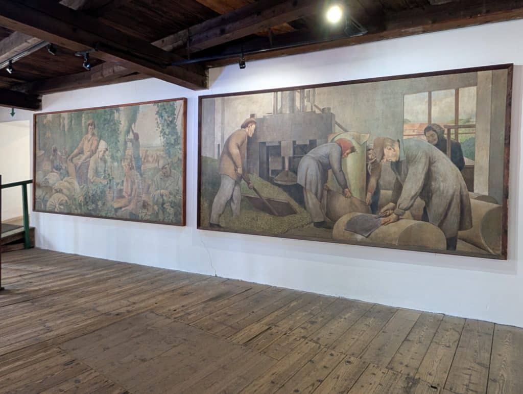 Zwei großformatige Gemälde im Hopfenmuseum Saaz zeigen in gedeckten Farben historische Szenen der Hopfenernte und der anschließenden Verarbeitung durch Arbeiter in einer Fabrikhalle.