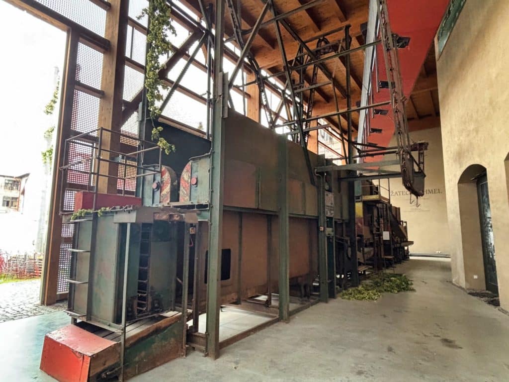 Eine große, historische Hopfenpflückmaschine aus Metall steht in einer hohen, holzverkleideten Halle des Hopfenmuseums in Žatec (Saaz). Das industrielle Gerät ist von einem komplexen Gerüst aus Stahlträgern und Förderbändern umgeben, an denen teilweise noch Hopfenreben hängen.