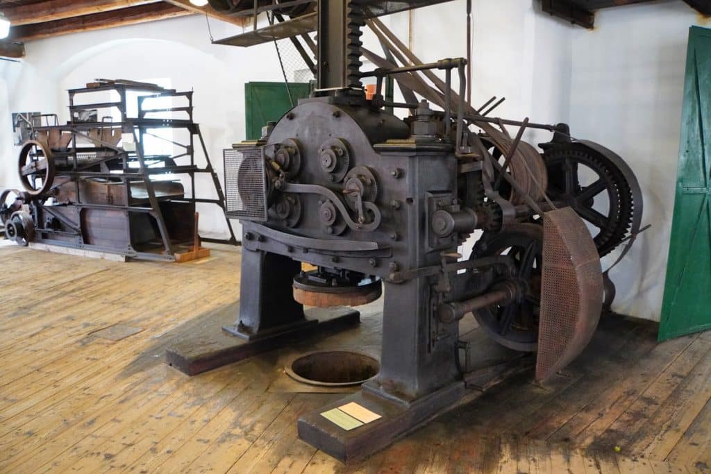 Eine historische Hopfenpresse aus massivem, dunklem Gusseisen steht im Zentrum des Saazer Hopfenmuseums auf einem rustikalen Dielenboden. Die komplexe Maschine mit Zahnrädern, Walzen und einem großen Pressstempel ist Teil einer Ausstellung alter landwirtschaftlicher Geräte.