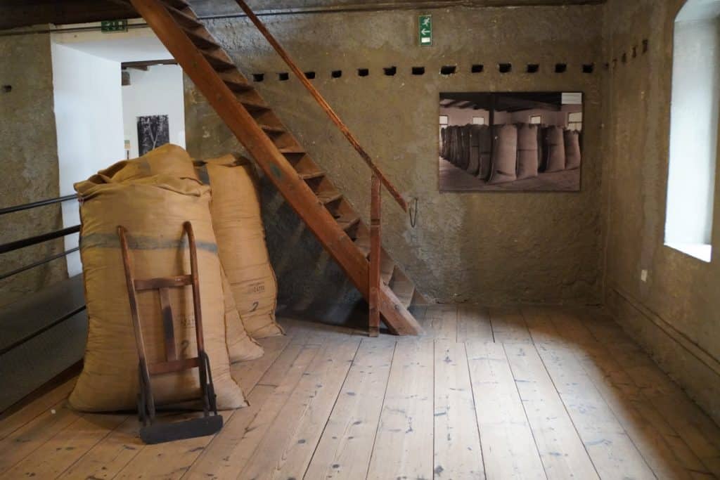 Ein Ausstellungsraum im Hopfenmuseum Saaz zeigt große, gefüllte Jutesäcke neben einer hölzernen Sackkarre und einer Treppe auf einem Dielenboden.