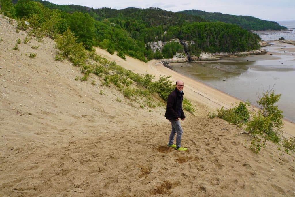 Ein Mann mit dunkler Jacke steigt eine steile Sanddüne in Tadoussac hinab, während sich im Hintergrund eine weite Bucht mit bewaldeten Hügeln erstreckt.