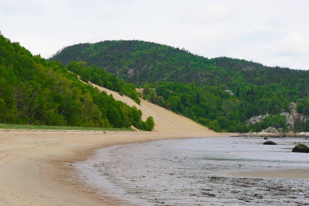 Ein weiter Sandstrand an der Bucht der Lorenzstroms führt zu einer hohen, hellen Sanddüne, die von dicht bewaldeten, grünen Hügeln unter einem bewölkten Himmel eingerahmt wird.