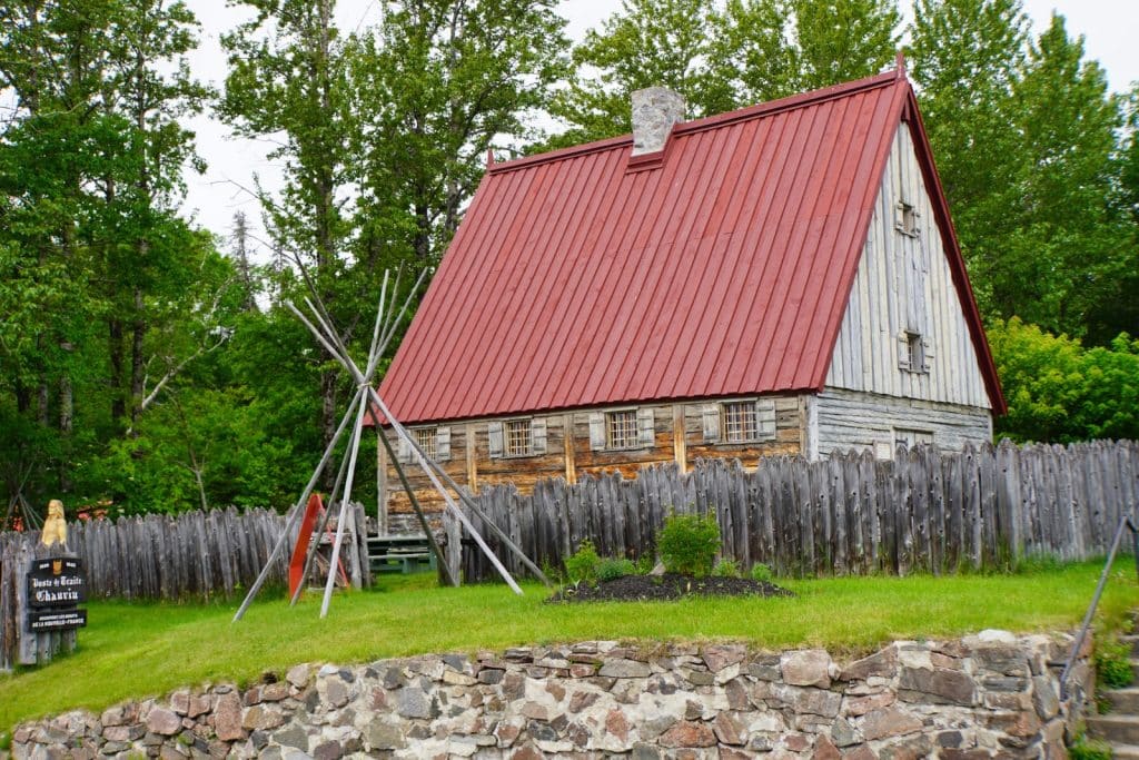 Tadoussac Sehenswürdigkeiten: Das historische Gebäude des Handelspostens mit seinem markanten roten Steildach und den Holzwänden steht hinter einem rustikalen Palisadenzaun, flankiert von einem hölzernen Tipi-Gerüst auf einer grünen Wiese.