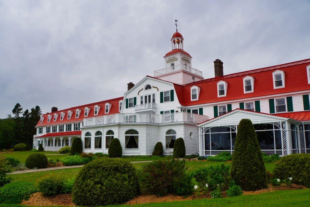 Tadoussac Sehenswürdigkeiten: Hotel Tadoussac mit seiner markanten weißen Fassade, dem leuchtend roten Dach und einem zentralen Glockenturm steht inmitten einer gepflegten Grünanlage unter bewölktem Himmel.