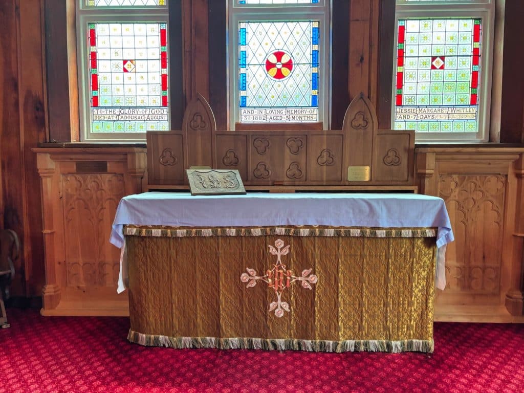 Ein hölzerner Altar bedeckt mit einem goldbestickten Tuch mit Kreuzmotiv, steht vor drei bunten Buntglasfenstern, die zum Gedenken an ein Kind gestiftet wurden.