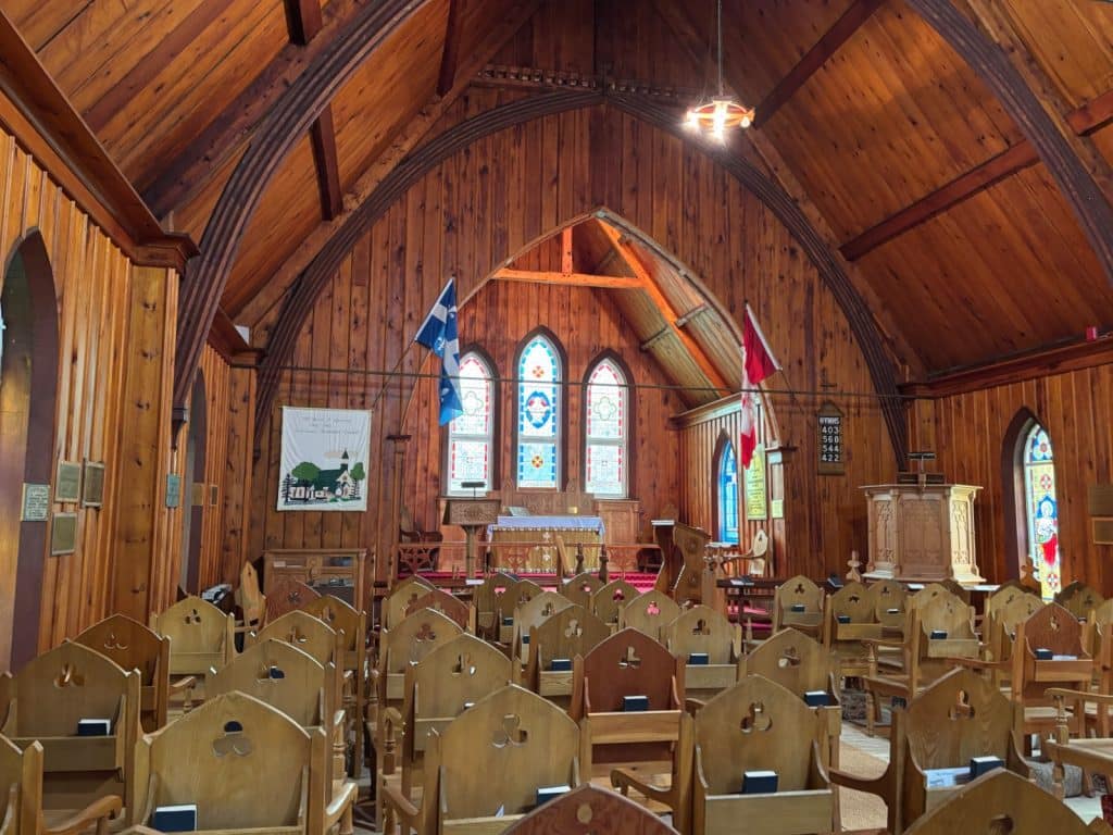 Das Innenraumfoto der hölzernen Kirche von Tadoussac zeigt ein warmes, rustikales Kirchenschiff mit spitzen Deckenbögen, hölzernen Stühlen und bunten Buntglasfenstern hinter dem Altar. Links und rechts hängen die Flaggen von Québec und Kanada, während die Wände und Decken vollständig mit vertikalen Holzpaneelen verkleidet sind.