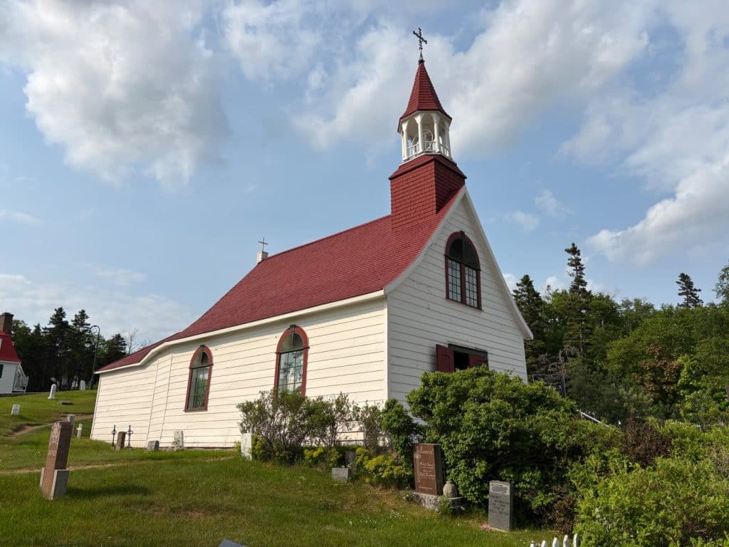 Tadoussac Sehenswürdigkeiten: Kirche St. Anna mit weißer Holzfassade, rotem Dach und einem kleinen Glockenturm steht auf einem grünen Hügel neben einem alten Friedhof unter blauem Himmel.