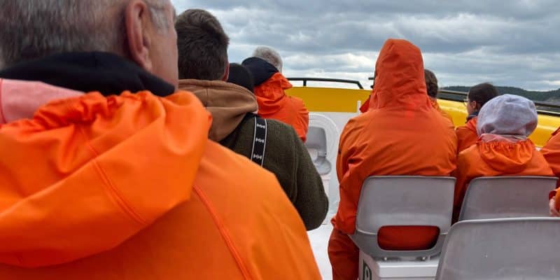 Passagiere in leuchtend orangefarbenen Regenjacken sitzen auf einem Zodiac-Boot während einer Waltour in Tadoussac unter einem bewölkten Himmel.