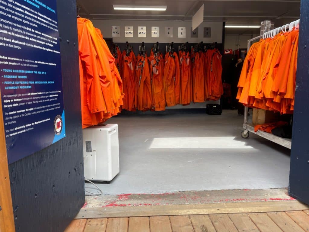 Blick in eine Kleiderkammer in der zahlreiche leuchtend orangefarbene Schutzanzüge nach Größen sortiert an den Wänden und auf Rollwagen hängen. Ein Hinweisschild mit Sicherheitsregeln ist im Vordergrund links an der blauen Tür angebracht.