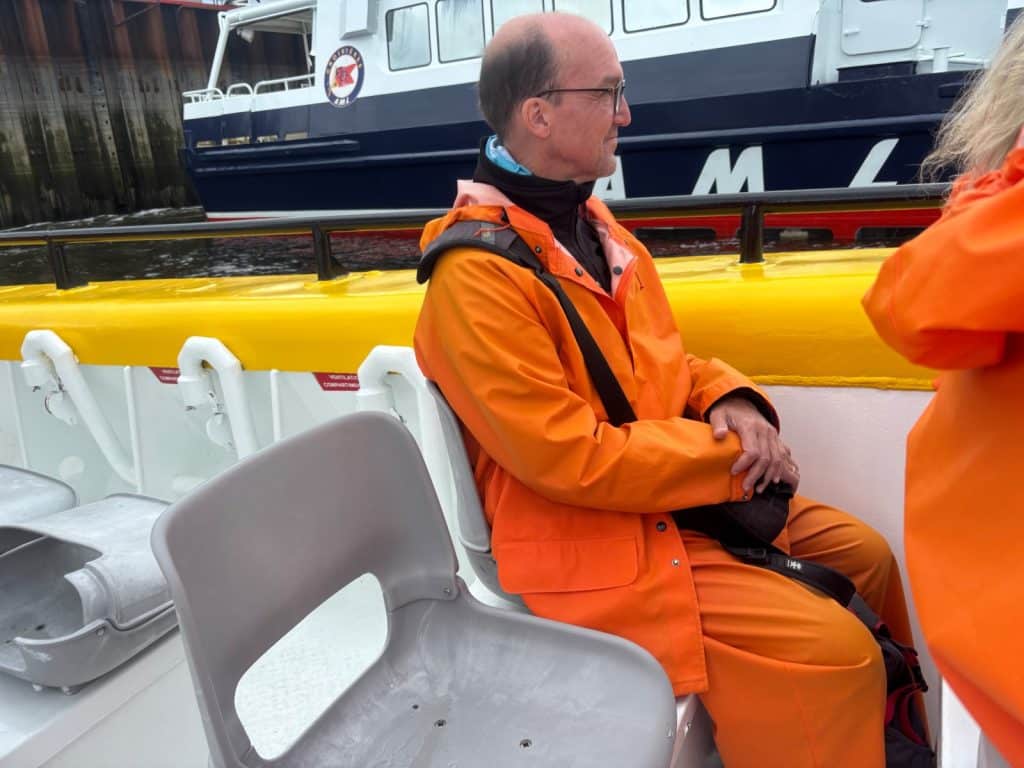 Ein Mann in einem leuchtend orangefarbenen Regenanzug sitzt zu Beginn einer Waltour in Tadoussac auf einem Boot und blickt auf das Wasser, während im Hintergrund ein weiteres Schiff vorbeifährt.