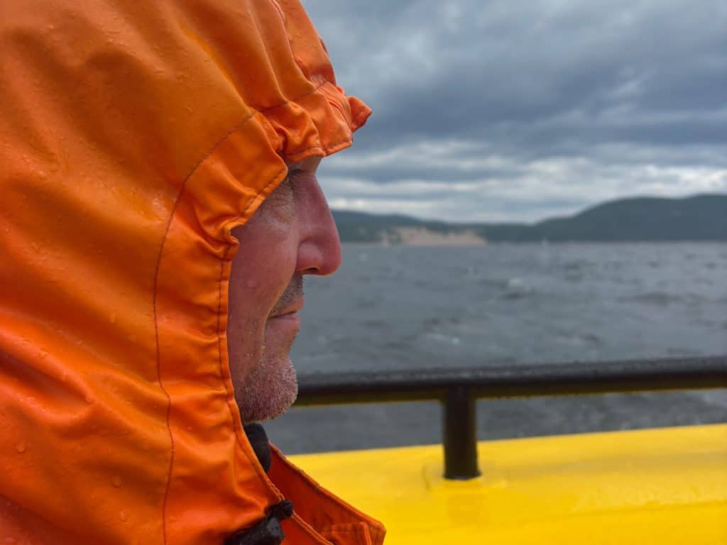 Nahaufnahme im Profil von Patrick, der während einer Waltour in Tadoussac eine leuchtend orangefarbene Regenjacke mit Kapuze trägt und auf das graue, wellige Meer blickt.