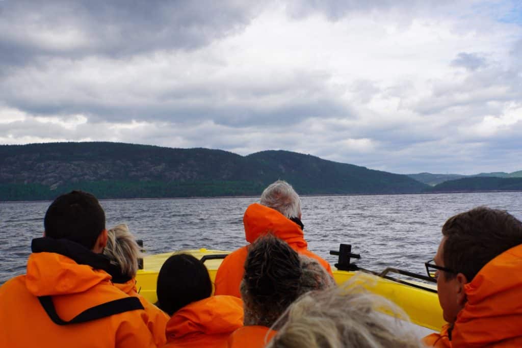 Eine Gruppe von Menschen in leuchtend orangefarbenen Regenjacken sitzt in einem gelben Boot auf dem Wasser vor Tadoussac und blickt während einer Waltour auf die bewaldeten Hügel am Ufer unter einem bewölkten Himmel.