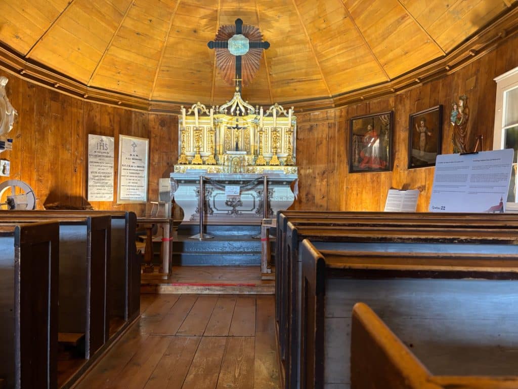 Innenansicht der hölzernen Kapelle mit Blick durch die Kirchenbänke auf den prunkvollen, goldverzierten Altar und ein großes Kruzifix an der Rückwand.