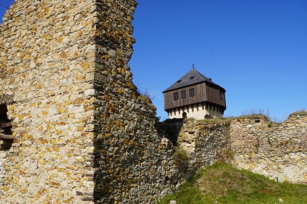 Die Ruinen der tschechischen Burg Hartenštejn zeigen eine zerfallene, helle Steinmauer im Vordergrund und einen gut erhaltenen Turm mit dunklem Holzoberbau unter einem strahlend blauen Himmel.