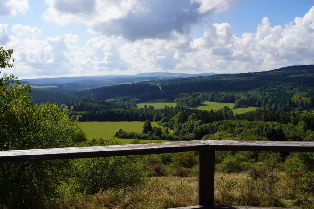 Blick von der Burg Hartenstein in Tschechien über eine weite, hügelige Landschaft mit grünen Wiesen und dichten Wäldern unter einem leicht bewölkten Himmel, eingerahmt von einem hölzernen Geländer im Vordergrund.