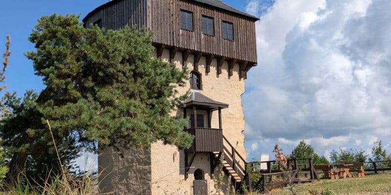 Der steinerne Aussichtsturm der Burg Hartenštejn in Tschechien verfügt über einen markanten hölzernen Oberbau mit Fenstern und ist über eine Außentreppe zugänglich. Er steht auf einer Anhöhe unter blauem Himmel mit weißen Wolken, flankiert von einer großen Kiefer und einer hölzernen Sitzgruppe.