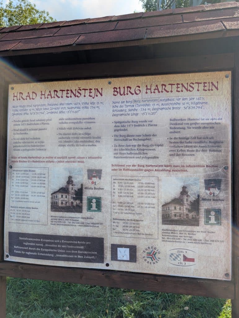 Ein hölzernes Hinweisschild in Tschechien informiert zweisprachig auf Tschechisch und Deutsch über die Geschichte und Besucherinformationen der Burg Hartenstein. Das Schild zeigt neben Textblöcken auch historische Abbildungen der Burg, das Wappen von Bochov sowie Kontaktinformationen und Öffnungszeiten.