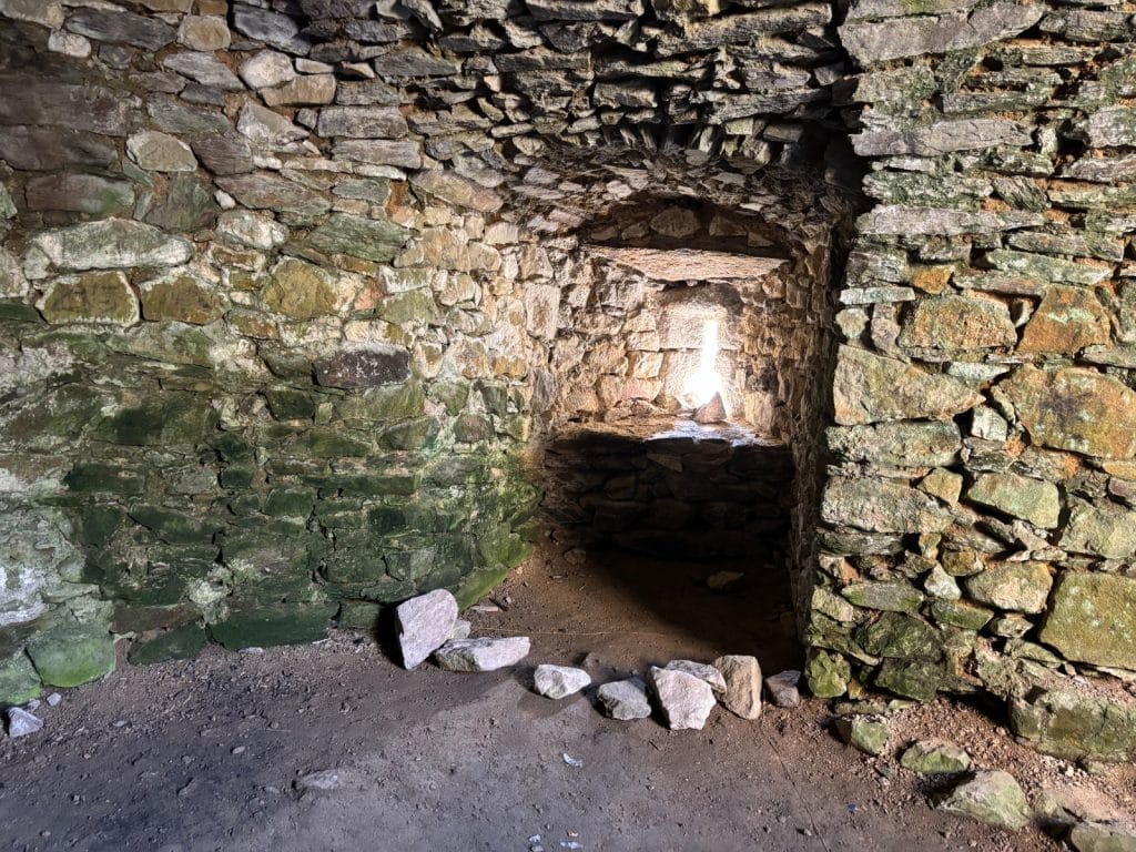 Ein Blick in den rustikalen Keller der Burg Hartenštejn in Tschechien, dessen Wände und gewölbte Decke aus groben, teils bemoosten Natursteinen gemauert sind und durch eine kleine Lichtöffnung im Hintergrund erhellt werden.