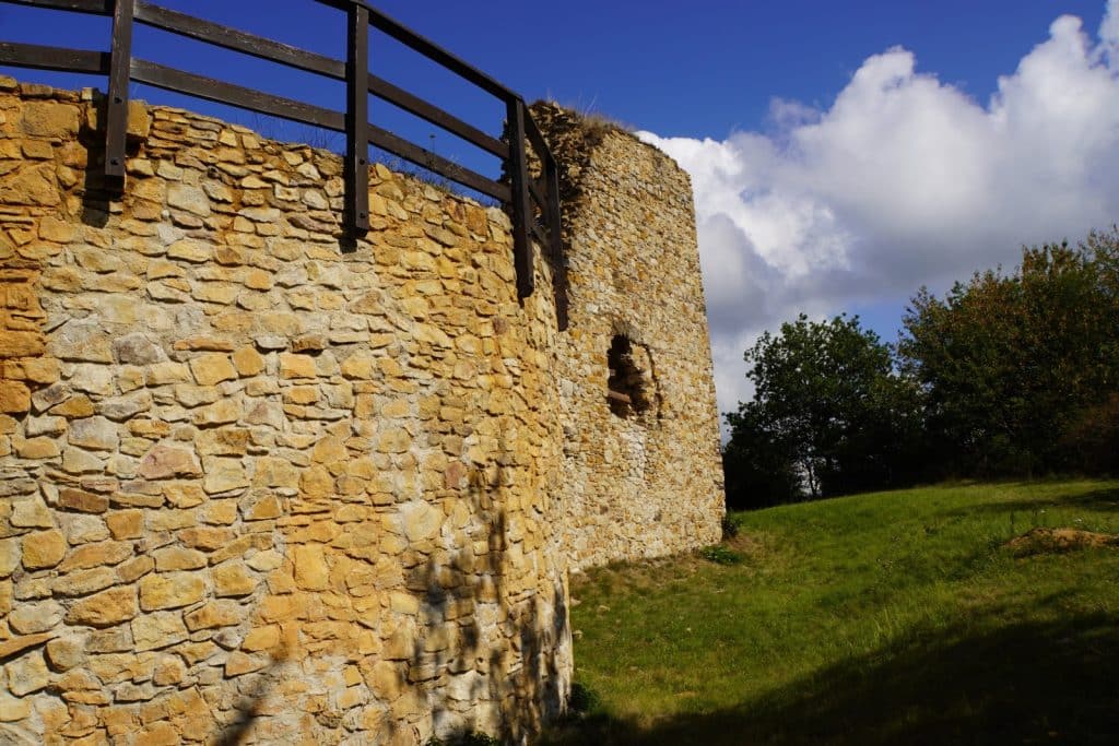 Die Ruinen der Burg Hartenštejn in Tschechien zeigen massive, hellbraune Steinmauern mit einem hölzernen Geländer an der Oberseite vor einem blauen Himmel mit weißen Wolken. Rechts erstreckt sich eine grüne Wiese mit Bäumen neben den verwitterten Überresten des historischen Mauerwerks.