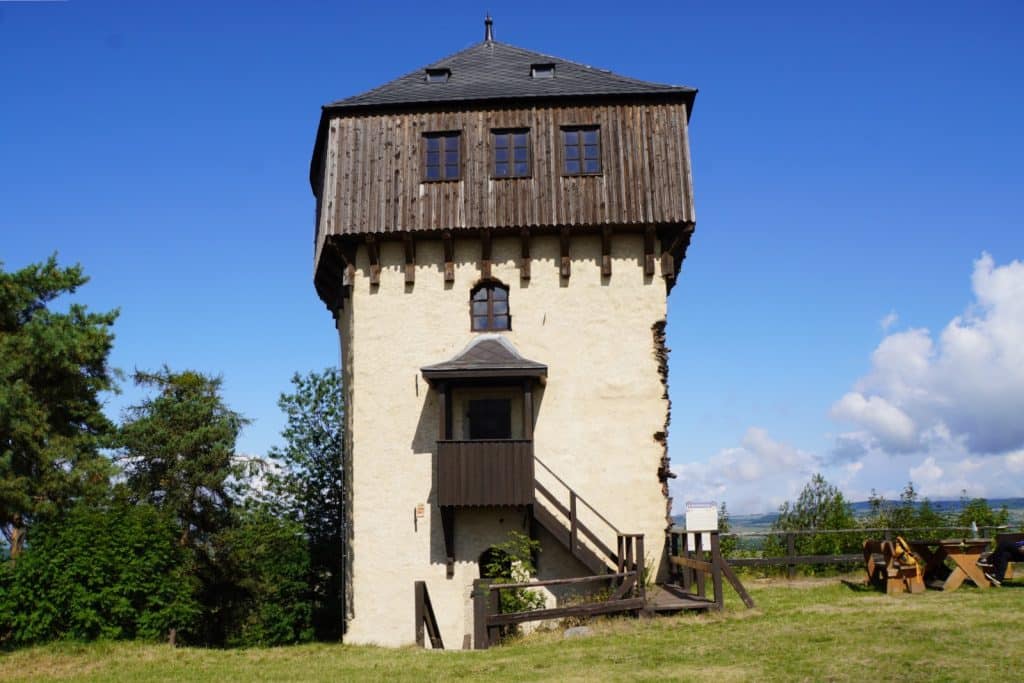 Der quadratische Turm der Burg Hartenštejn in Tschechien mit einer hellen Steinfassade, einem hölzernen Obergeschoss und einem überdachten Außeneingang steht unter strahlend blauem Himmel auf einer Wiese.