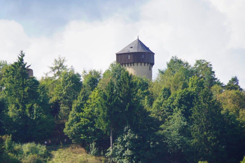 Blick aus der Ferne auf den hölzernen Turm der tschechischen Burg Hartenštejn, der über die dichten Baumkronen eines bewaldeten Hügels ragt.