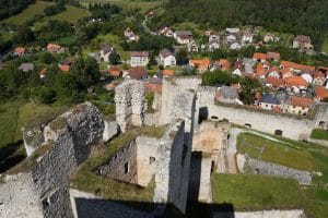 Blick von oben auf die steinernen Ruinen der Burg Rabí in Tschechien, die im Hintergrund an ein Dorf mit rot gedeckten Häusern und grüne Hügel grenzen.