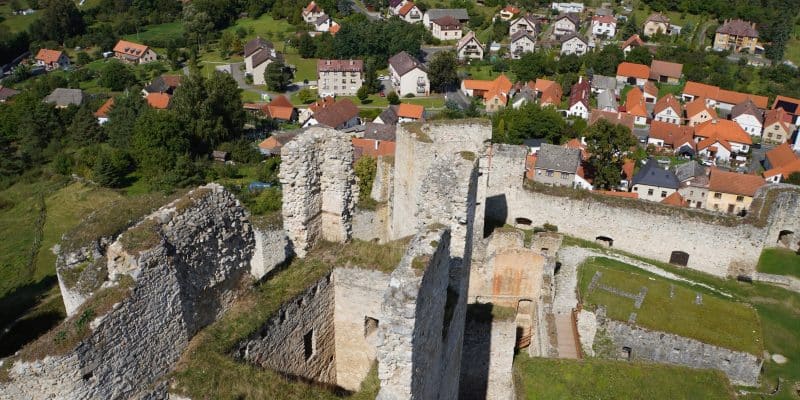 Blick von oben auf die steinernen Ruinen der Burg Rabí in Tschechien, die im Hintergrund an ein Dorf mit rot gedeckten Häusern und grüne Hügel grenzen.