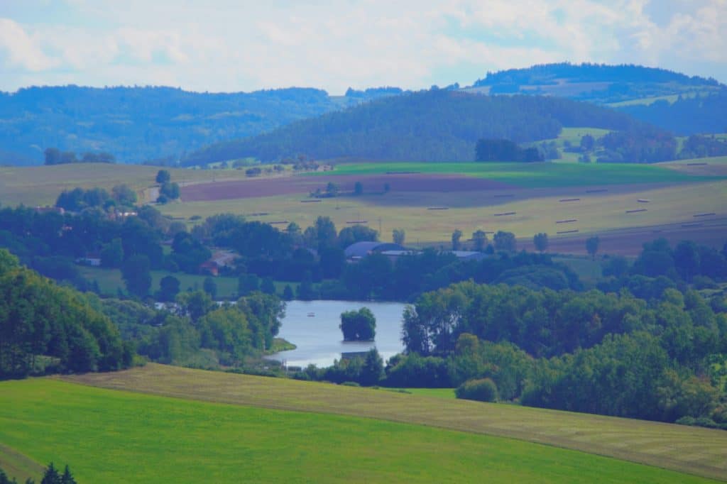 Ein weiter Ausblick von der Burg Rabí in Tschechien über eine hügelige, grün-blaue Landschaft mit Feldern, Wäldern und einem kleinen See mit einer bewaldeten Insel im Zentrum. Im Hintergrund erstrecken sich sanfte, bewaldete Hügelketten unter einem leicht bewölkten Himmel.
