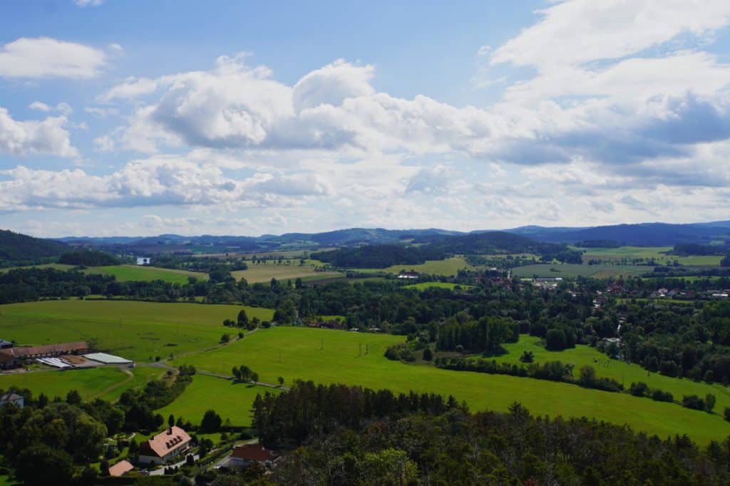Weite Panoramaaussicht von der Burg Rabí auf die hügelige tschechische Landschaft mit grünen Feldern, Wäldern und vereinzelten Häusern unter einem leicht bewölkten blauen Himmel.