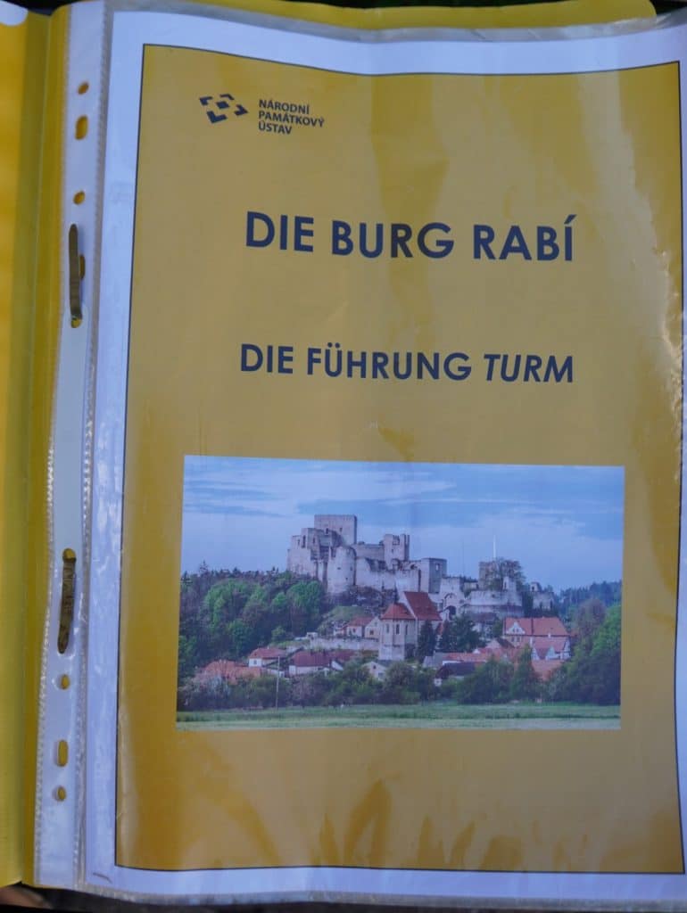 Ein Foto zeigt das Deckblatt einer deutschsprachigen Broschüre mit dem Titel „Die Führung Turm“, herausgegeben vom Nationalen Denkmalinstitut (Národní památkový ústav). Unter dem Text ist eine Panoramaaufnahme der weitläufigen Burganlage auf einem Hügel über einer kleinen Ortschaft abgebildet.