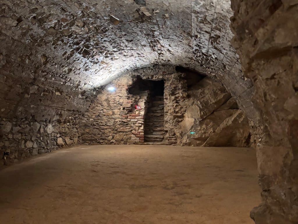 Ein weitläufiges, unterirdisches Kellergewölbe der Burg Rabí in Tschechien mit groben Steinwänden, einem sandigen Boden und einer schmalen Steintreppe, die in einen dunklen Gang führt.