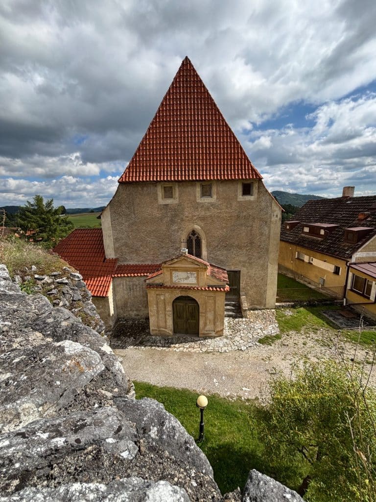Blick von einer Steinmauer hinab auf die mittelalterliche Kirche der Burg Rabí in Tschechien, die sich durch ein hohes, spitzes Ziegeldach und ein kleines barockes Eingangsportal auszeichnet.