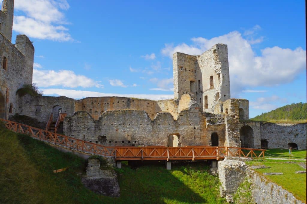 Die Ruinen der Seitengebäude der tschechischen Burg Rabí zeigen verwitterte Steinmauern und einen markanten Turm unter blauem Himmel, erschlossen durch einen hölzernen Steg im Vordergrund.