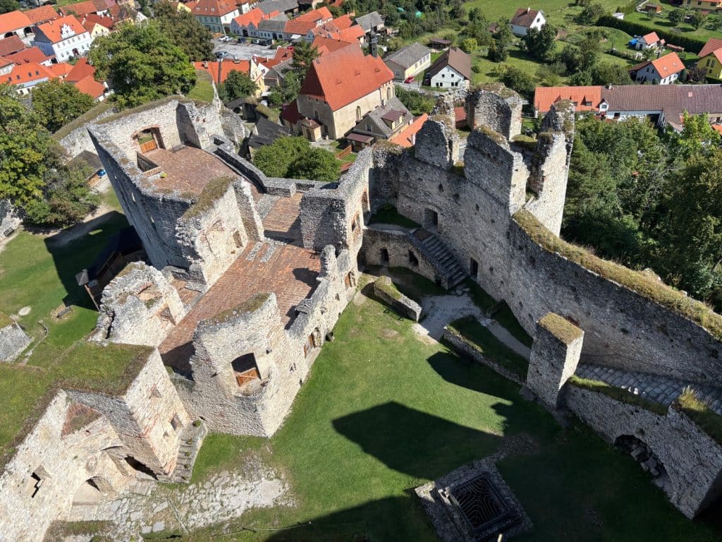 Blick von oben auf die steinernen Ruinen der Burg Rabí in Tschechien mit ihren grasbewachsenen Innenhöfen, umgeben von einem Dorf mit rot gedeckten Häusern.