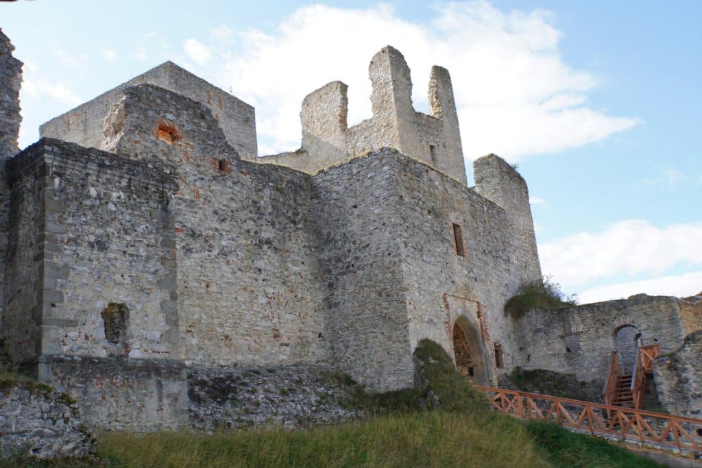 Die massiven, verwitterten Steinmauern und Ruinen der Burg Rabí in Tschechien ragen unter einem leicht bewölkten Himmel empor, wobei ein hölzerner Steg zum bogenförmigen Zugang des inneren Burghofs führt.