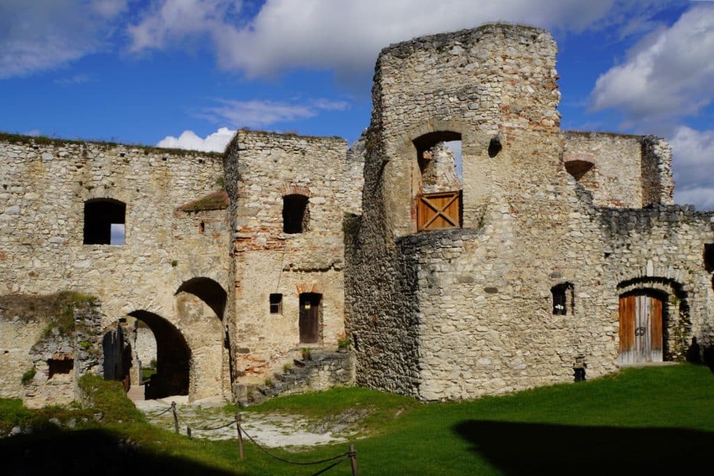Blick in den sonnigen zweiten Burghof der Burg Rabí in Tschechien mit seinen massiven, teils verfallenen Steinmauern, Torbögen und hölzernen Türen unter blauem Himmel.